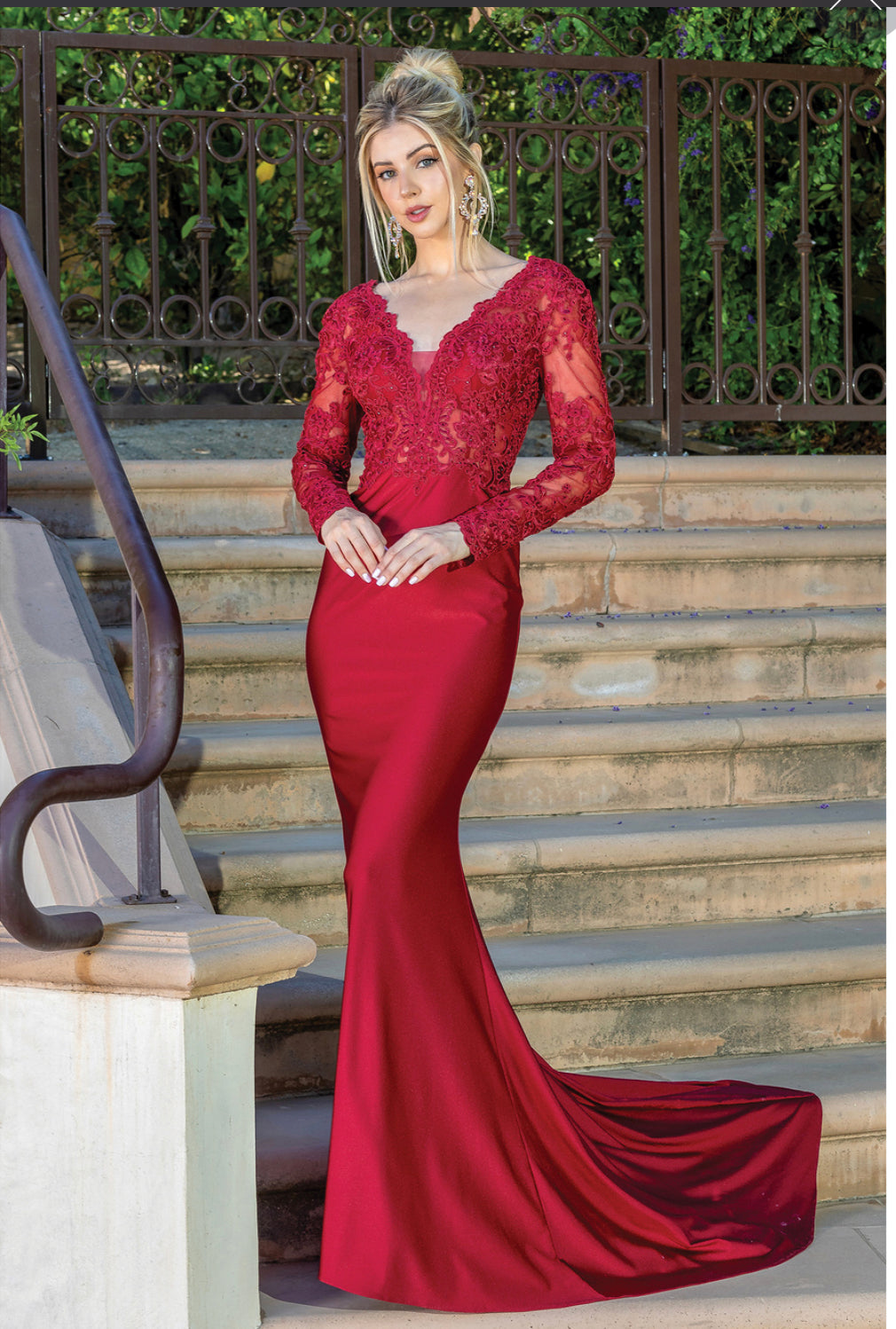 A woman stands on outdoor steps near a railing wearing the Long Sleeve Long Formal Prom Dress with Lace 4124—a fitted red evening gown featuring lace sleeves and a mermaid-style train.